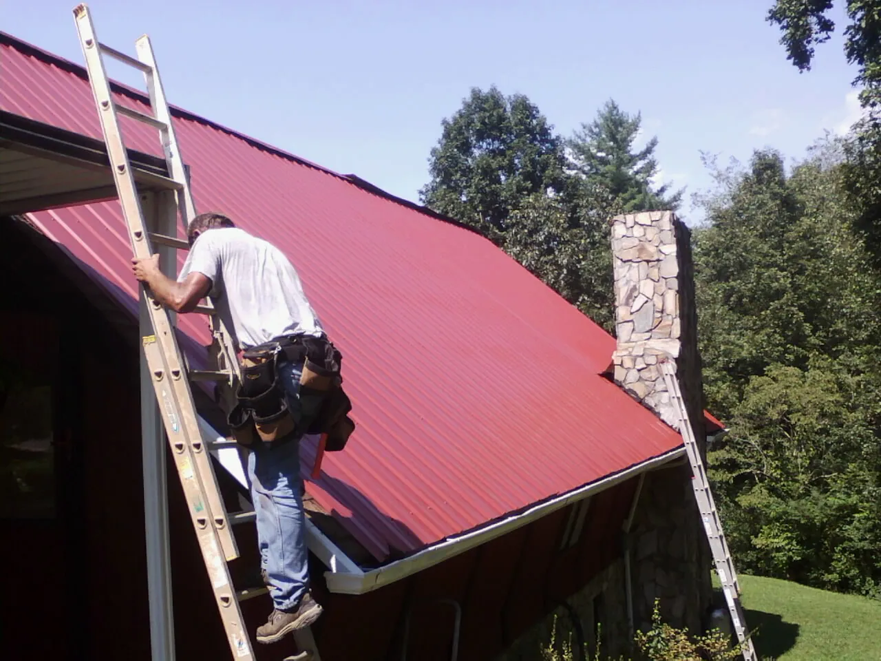 Roofer on a ladder beside a red metal roof and stone chimney — Soffit & Fascia Repair by Roofing Brothers in Kenedy